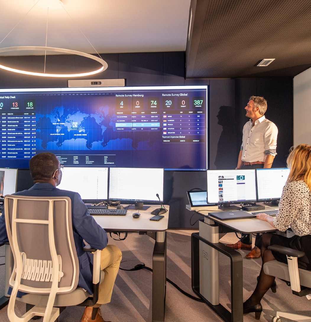 A control room, three people, two sitting one standing. Large screens and computers on desks.